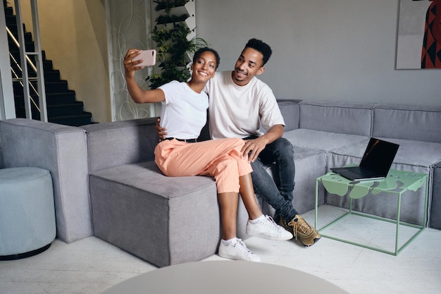 Jovem lindo casal afro-americano casual feliz tomando selfie no celular juntos no sofá em casa moderna