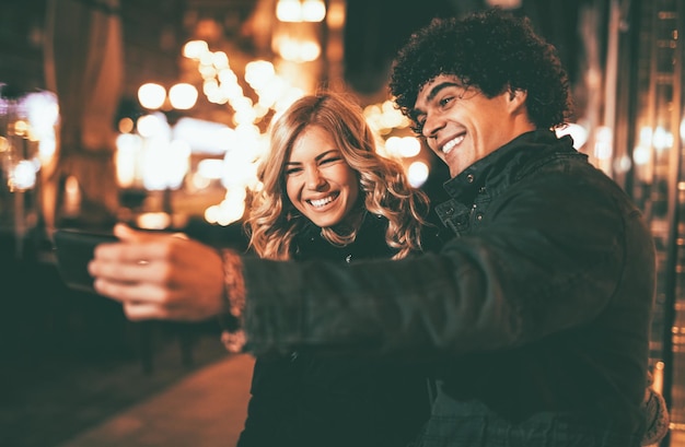 Jovem lindo casal alegre tomando uma selfie na rua da cidade na noite de ano novo com muitas luzes no fundo.