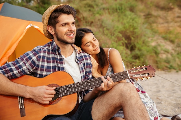 Jovem lindo casal feliz tocando violão enquanto está sentado na praia