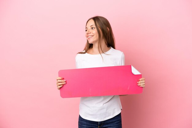 Foto jovem lituana isolada em fundo rosa segurando um cartaz vazio e olhando de lado