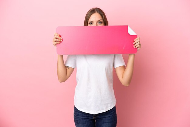 Jovem lituana isolada em fundo rosa segurando um cartaz vazio e se escondendo atrás dele