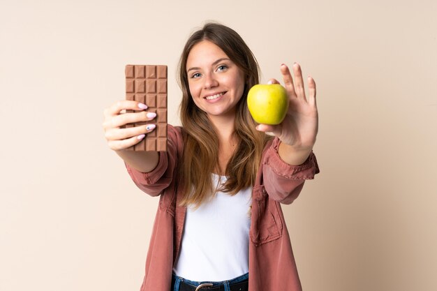 Jovem lituana isolada em um fundo bege pegando um tablete de chocolate em uma das mãos e uma maçã na outra