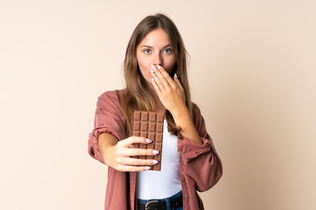 Foto jovem lituana isolada em um fundo bege tomando uma pastilha de chocolate e surpresa