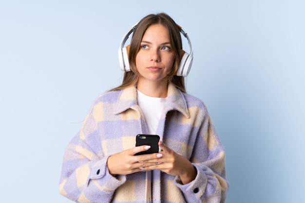 Jovem lituana isolada na parede azul ouvindo música com um celular e pensando