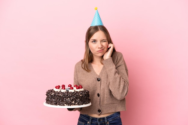 Jovem lituana segurando bolo de aniversário isolado em fundo rosa frustrado e cobrindo os ouvidos