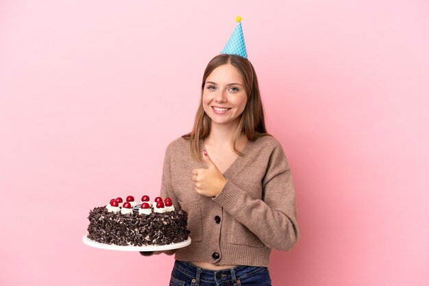 Jovem lituana segurando um bolo de aniversário isolado em um fundo rosa e fazendo um gesto de polegar para cima