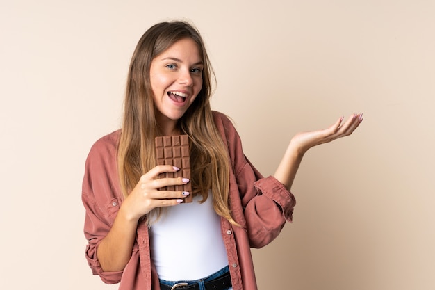 Jovem lituana surpresa isolada em uma parede bege tomando um comprimido de chocolate