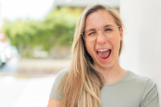 Foto jovem loira ao ar livre com óculos e expressão feliz