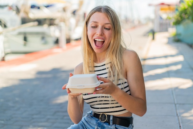 Jovem loira ao ar livre levando uma caixa de comida para viagem com expressão de surpresa