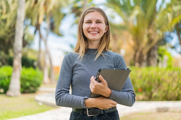 Jovem loira ao ar livre segurando um tablet com expressão feliz