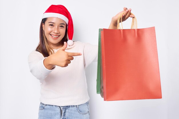 Jovem loira asiática sorridente de 20 anos usando um chapéu de Natal e apontando o dedo para sacolas de compras depois de comprar acessórios de Natal isolados no fundo branco Feliz Ano Novo Natal 2023