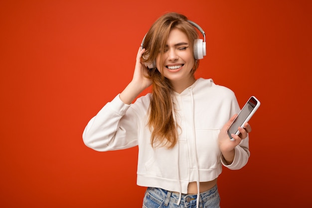 Jovem loira atraente e feliz usando um capuz branco isolado sobre uma parede de fundo colorido