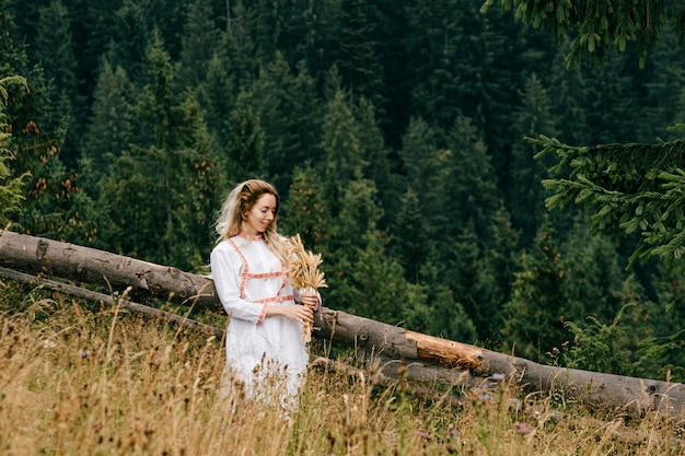 Jovem loira atraente em um vestido branco com bordado posando com buquê de espigas em uma paisagem pitoresca.