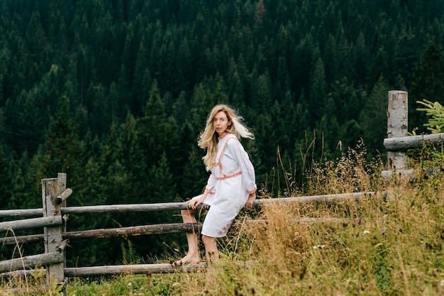 Jovem loira atraente em um vestido branco com bordado sentada na cerca de madeira sobre a pitoresca paisagem da floresta