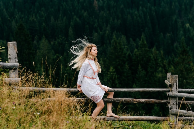 Jovem loira atraente em um vestido branco com bordado sentada na cerca de madeira sobre a pitoresca paisagem da floresta