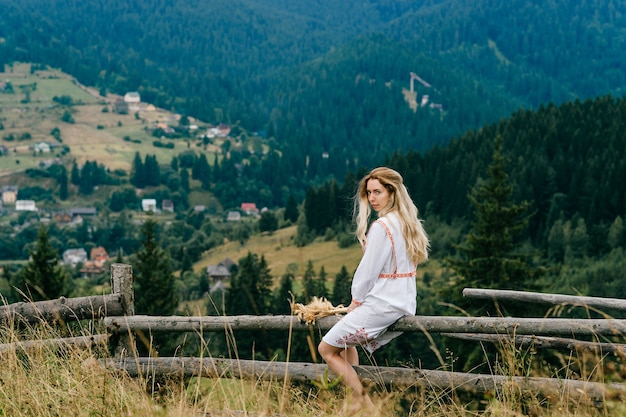 Jovem loira atraente em um vestido branco com enfeite sentado na cerca de madeira com buquê de espigas sobre a pitoresca paisagem rural