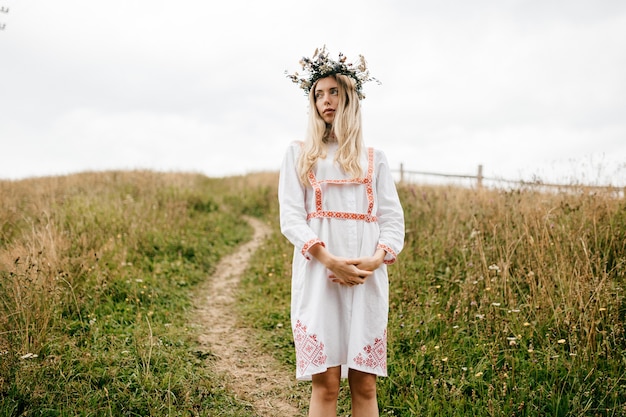 Jovem loira atraente em um vestido branco com ornamento e guirlanda de flores na cabeça posando no campo