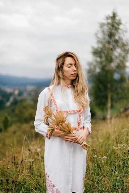 Jovem loira atraente em um vestido branco com ornamento posando com buquê de espigas sobre a pitoresca paisagem rural