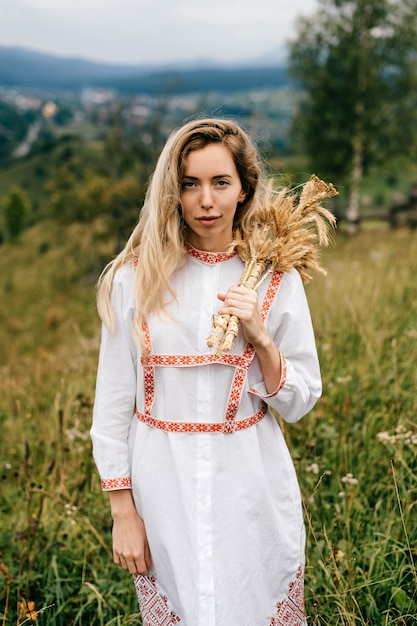 Jovem loira atraente em um vestido branco com ornamento posando com buquê de espigas sobre o fundo do campo