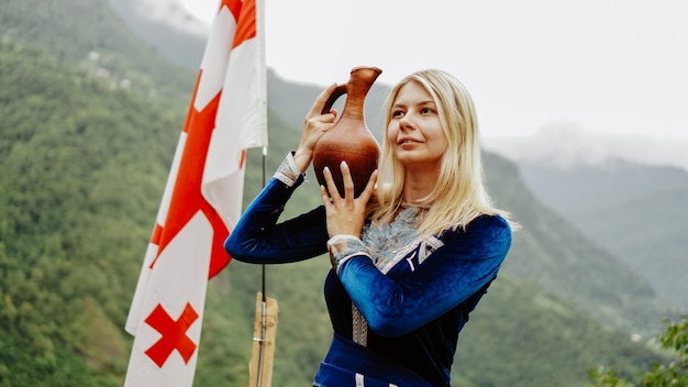 Jovem loira bonita com roupas da Geórgia no contexto da bandeira da Geórgia e as montanhas