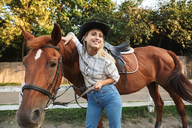 Jovem loira bonita e sorridente acariciando um cavalo no pátio dos cavalos