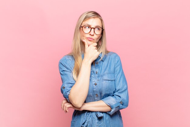 Jovem loira bonita parecendo séria, pensativa e desconfiada, com um braço cruzado e a mão no queixo, opções de ponderação