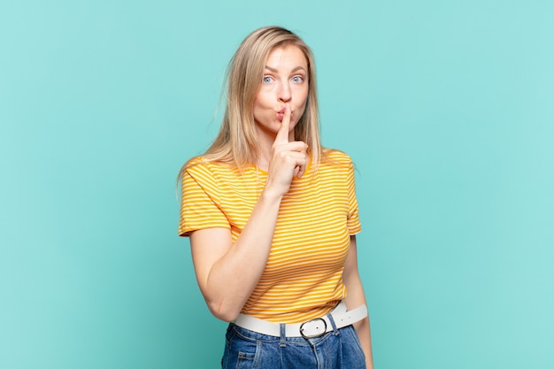 Jovem loira bonita pedindo silêncio e silêncio, gesticulando com o dedo na frente da boca, dizendo shh ou guardando segredo