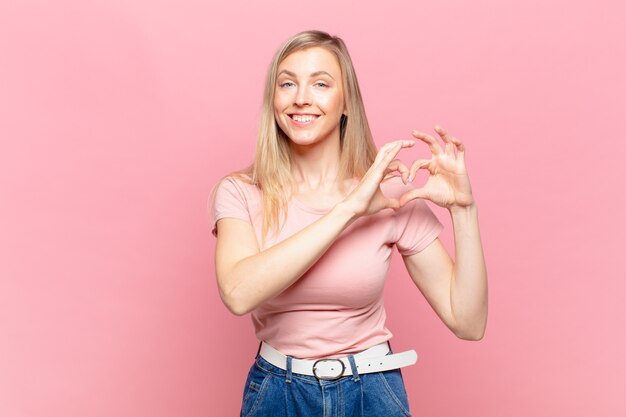 jovem loira bonita sorrindo e se sentindo feliz, fofa, romântica e apaixonada, fazendo formato de coração com as duas mãos