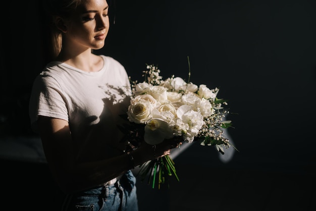 Jovem loira bonita vestindo roupas de moda segurando a composição maravilhosa de flores frescas nas mãos sobre fundo branco, olhando para a câmera.