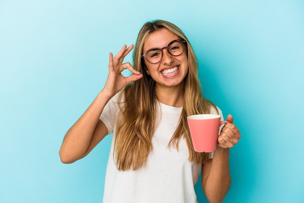 Jovem loira branca segurando uma caneca isolada na parede azul alegre e confiante mostrando um gesto de ok