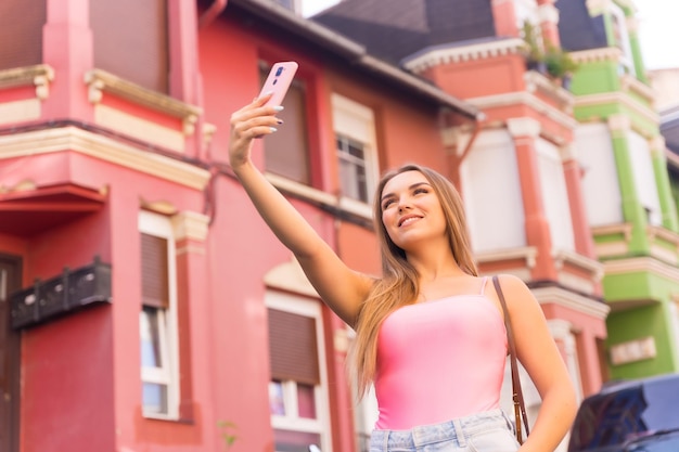 Jovem loira caucasiana em uma rua com casas com fachadas coloridas sorrindo tirando uma selfie