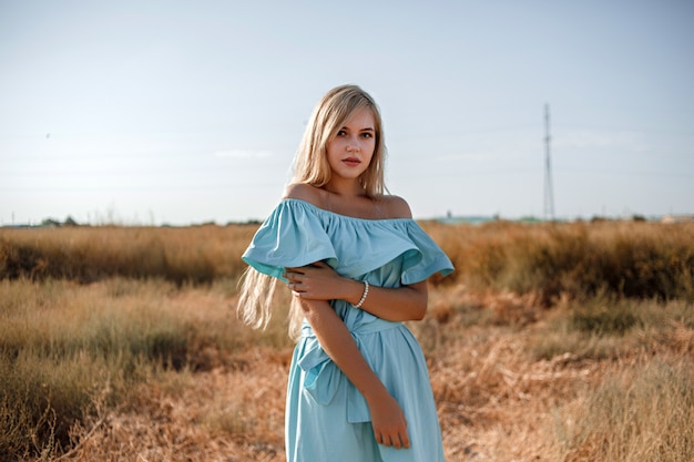 Jovem loira caucasiana linda num vestido azul claro fica no campo com a grama queimada pelo sol