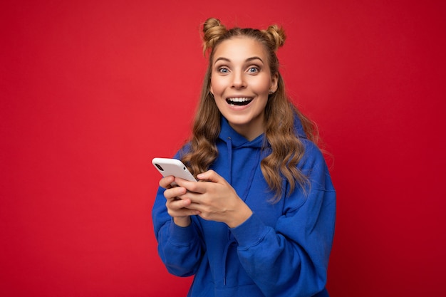 Jovem loira chocada e atraente com capuz azul e elegante, isolada sobre fundo vermelho, enviando mensagens de texto via telefone, olhando para a câmera