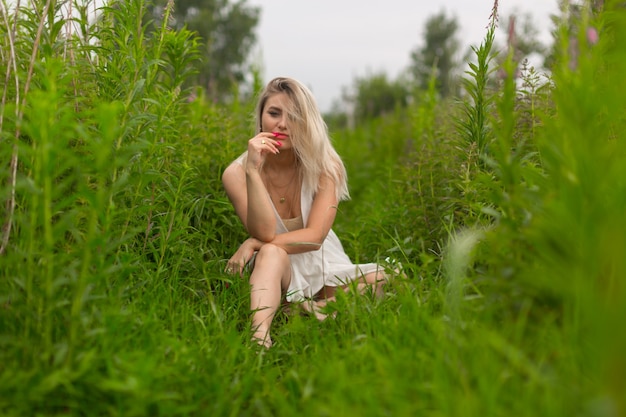Jovem loira com batom vermelho e vestido de verão senta-se entre a grama alta e verde.