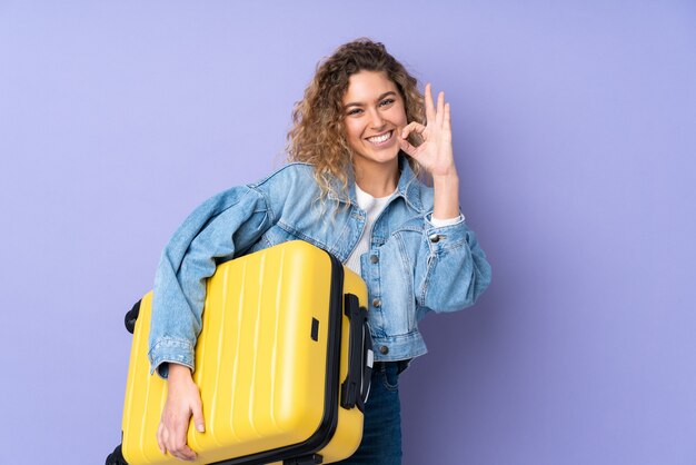 Jovem loira com cabelo encaracolado isolado na parede roxa nas férias com mala de viagem e fazendo sinal de OK