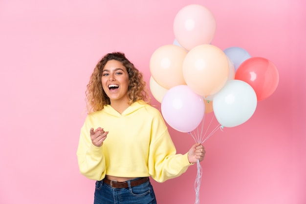 Jovem loira com cabelo encaracolado pegando muitos balões isolados na parede rosa aponta o dedo para você