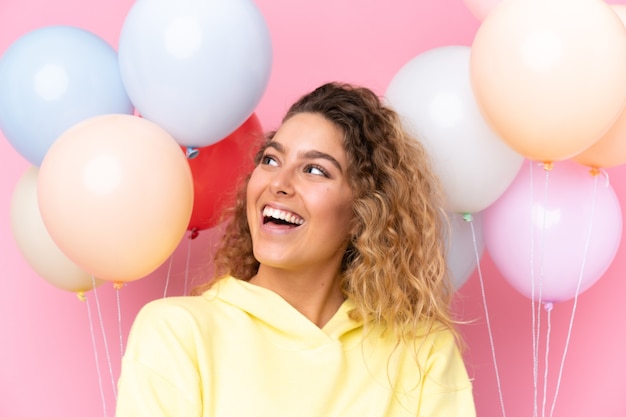 Jovem loira com cabelo encaracolado pegando muitos balões isolados na parede rosa