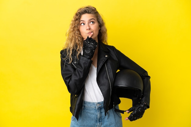 Jovem loira com capacete de motociclista isolada em um fundo amarelo tendo dúvidas enquanto olha para cima