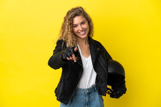 Foto jovem loira com um capacete de motociclista isolado em um fundo amarelo apontando para a frente com uma expressão feliz