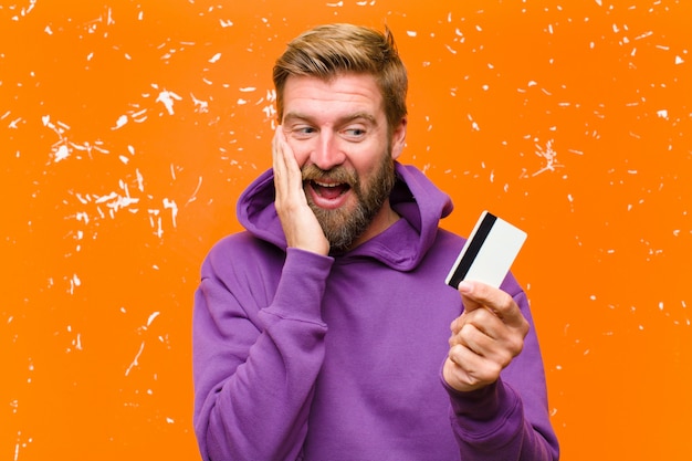 Jovem loira com um cartão de crédito, vestindo um capuz roxo contra parede laranja danificada