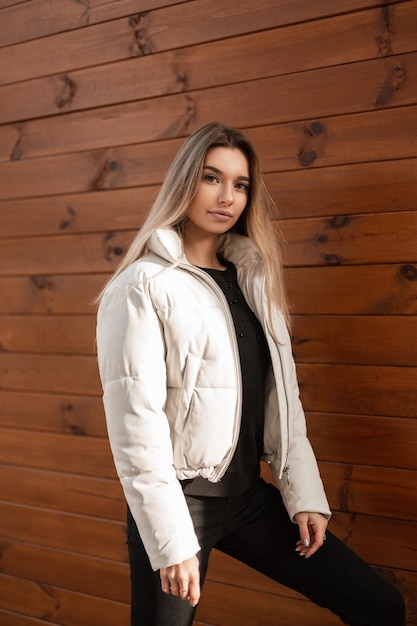 Jovem loira com um suéter preto e uma elegante jaqueta branca posando perto de um prédio de madeira