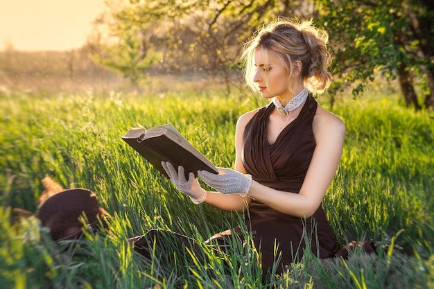 Foto jovem loira com um vestido vintage marrom e cartola lendo um livro no jardim primavera