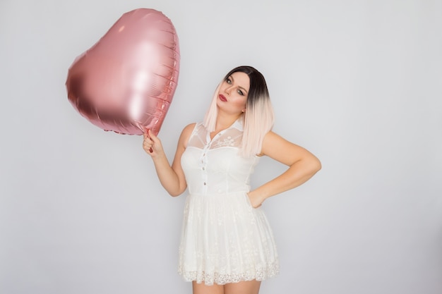 Jovem loira com vestido de renda branca segurando um grande balão de coração rosa nas mãos sobre fundo branco