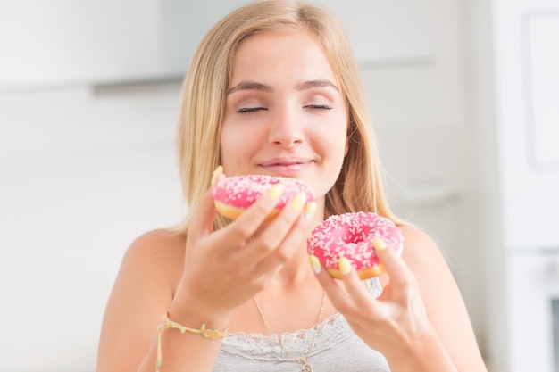 Jovem loira come rosquinhas rosa na cozinha de casa com emoções de sabor.