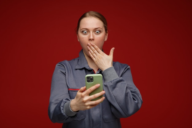 Jovem loira confusa, engenheira, vestindo uniforme, segurando e olhando para o celular, fazendo gesto de opa