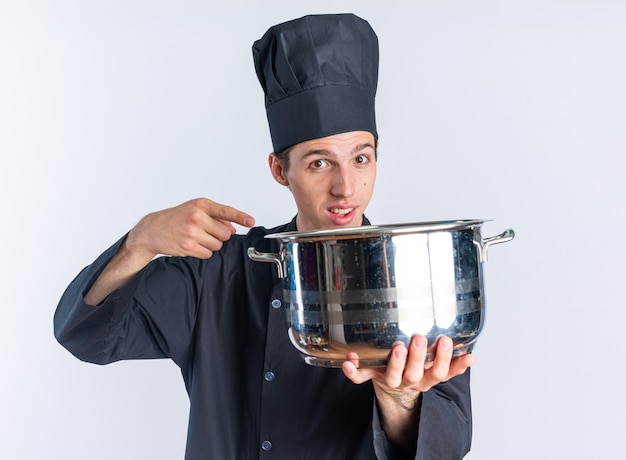 Jovem loira cozinheira impressionada com uniforme de chef e boné estendendo a panela em direção à câmera, apontando para ela, olhando para a câmera isolada na parede branca