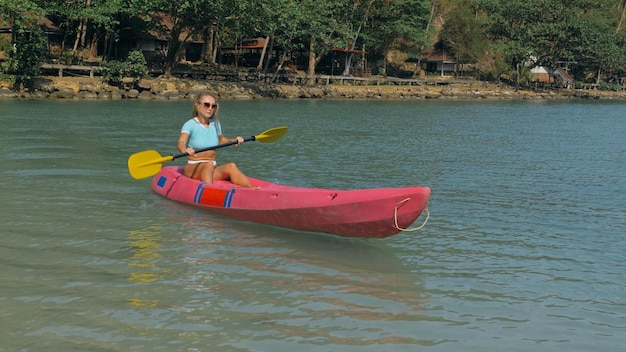 Foto jovem loira de maiô azul enfileira canoa de plástico rosa ao longo da baía do mar azul