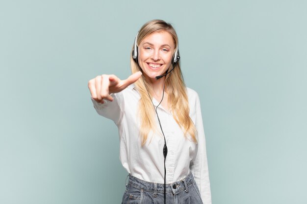 Jovem loira de telemarketing apontando para a câmera com um sorriso satisfeito, confiante e amigável, escolhendo você