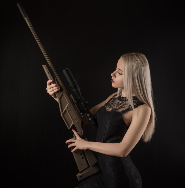Jovem loira de vestido preto posando em um fundo preto com uma arma
