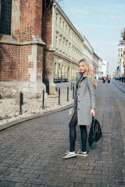 Foto jovem loira e bonita com cabelos ondulados loiros compridos andando na rua e segurando a mochila e o buquê.